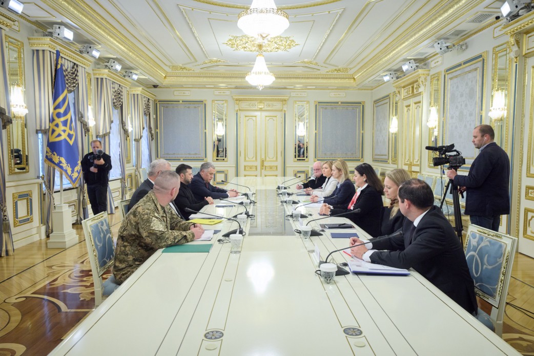 Дві групи офіційних осіб у цивільному та військовому одязі проводять зустріч за довгим столом у розкішній залі з класичним інтер’єром, кришталевими люстрами та позолотою. У куті зали встановлено державний прапор України, а подію фіксують оператори з відеокамерами.