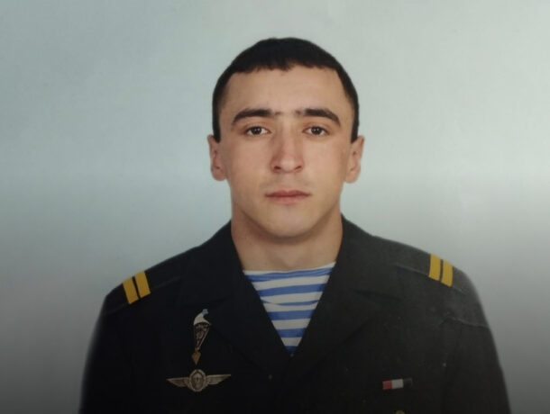 Портретний знімок молодого чоловіка зі стрижкою під нуль та серйозним виразом обличчя, одягненого у темно-синю військову форму з погонами та значком парашутиста на грудях поверх тільника у синю смужку.