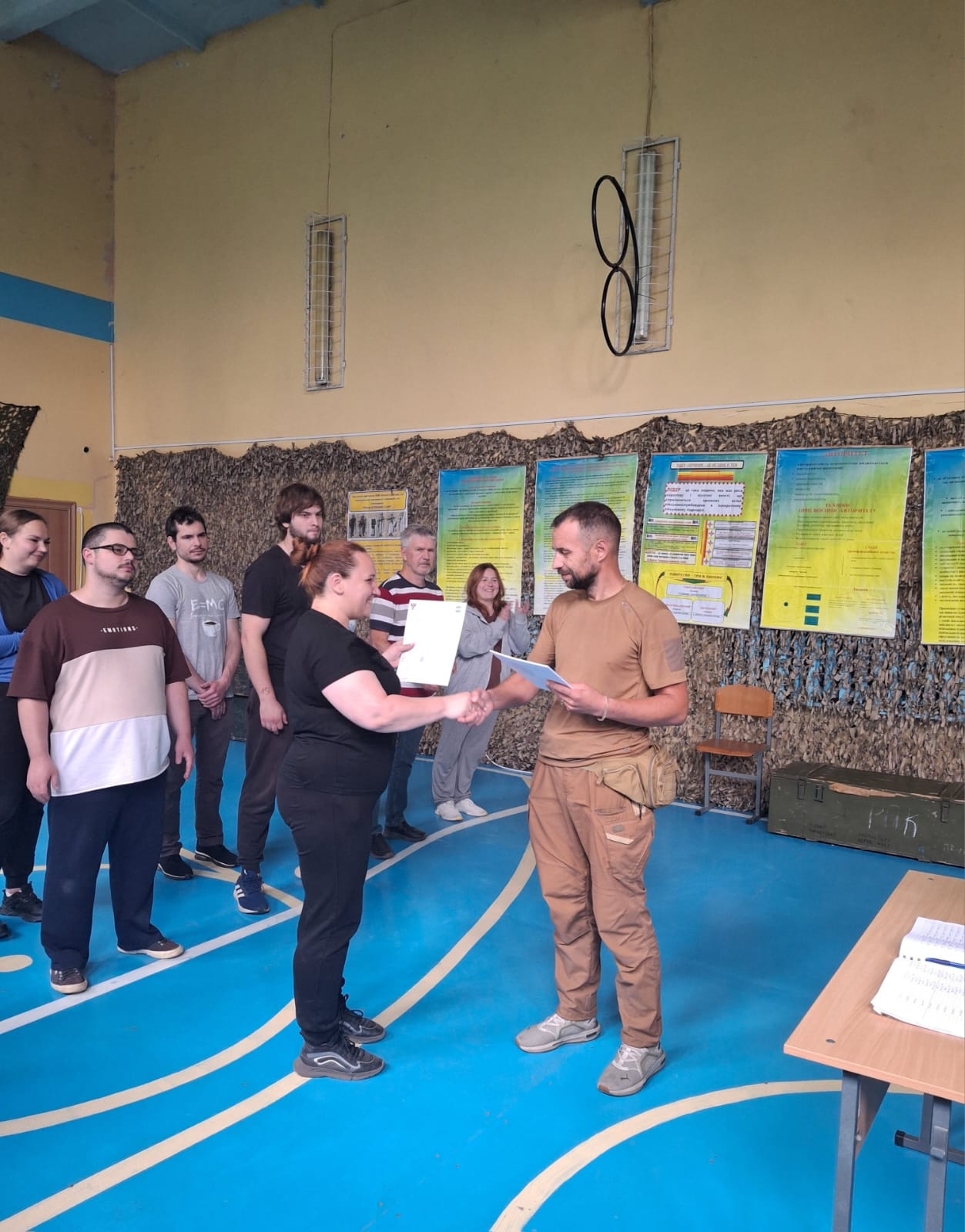 Урочисте вручення сертифікатів після завершення навчального курсу з підготовки. Інструктор у тактичному пісочному одязі та з поясною сумкою тисне руку жінці в чорному одязі та передає їй сертифікат про навчання. Позаду стоїть шеренга інших учасників курсу, які спостерігають та аплодують. Захід проходить у спортивній залі, де стіни задекоровані маскувальною сіткою, навчальними плакатами, а в кутку стоїть військовий ящик для боєприпасів.