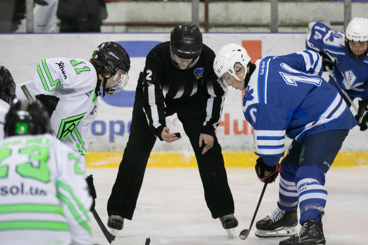 Суддя готується до вкидання шайби на льодовій арені під час хокейного матчу. Два гравці у біло-зеленій та синьо-білій формі схилилися один навпроти одного, тримаючи ключки напоготові.