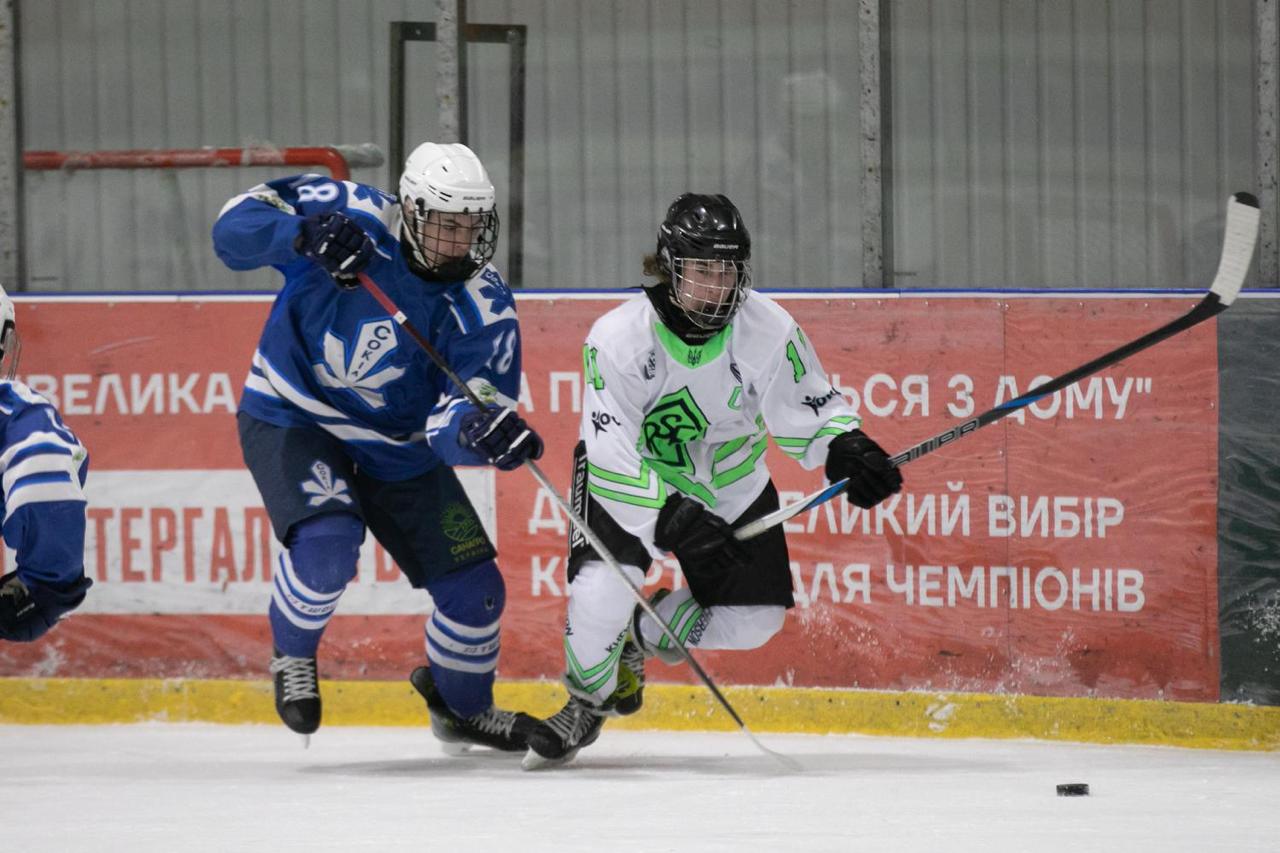 Динамічний момент хокейного матчу на льодовій арені. Два хокеїсти у синій та біло-зеленій формі ведуть активну боротьбу за шайбу. На фоні видно захисний борт із рекламними написами українською мовою.