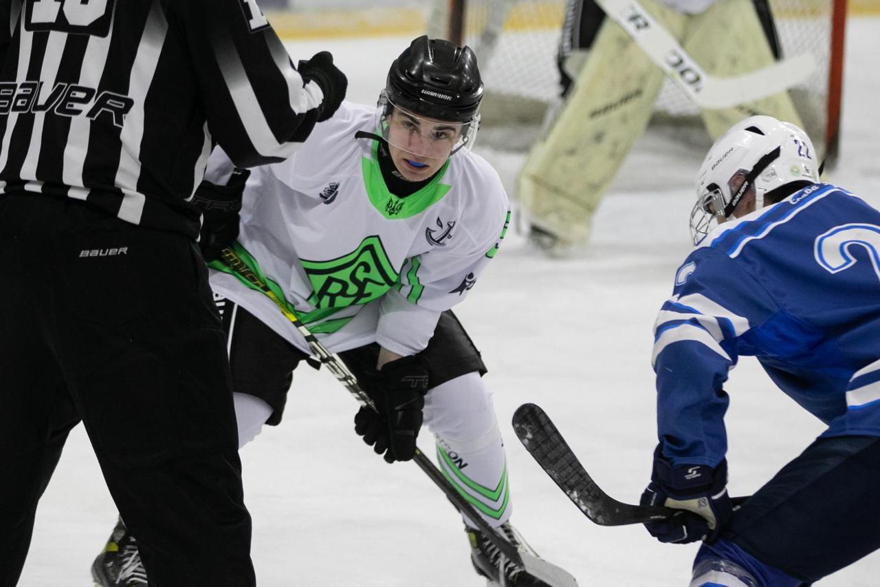 Хокеїст у білій формі з яскраво-зеленими елементами зосереджено готується до вкидання шайби, тримаючи ключку на льоду. Навпроти нього перебуває гравець у синій формі, а поруч стоїть арбітр у класичному смугастому джерсі. На задньому плані видно ворота та голкіпера.