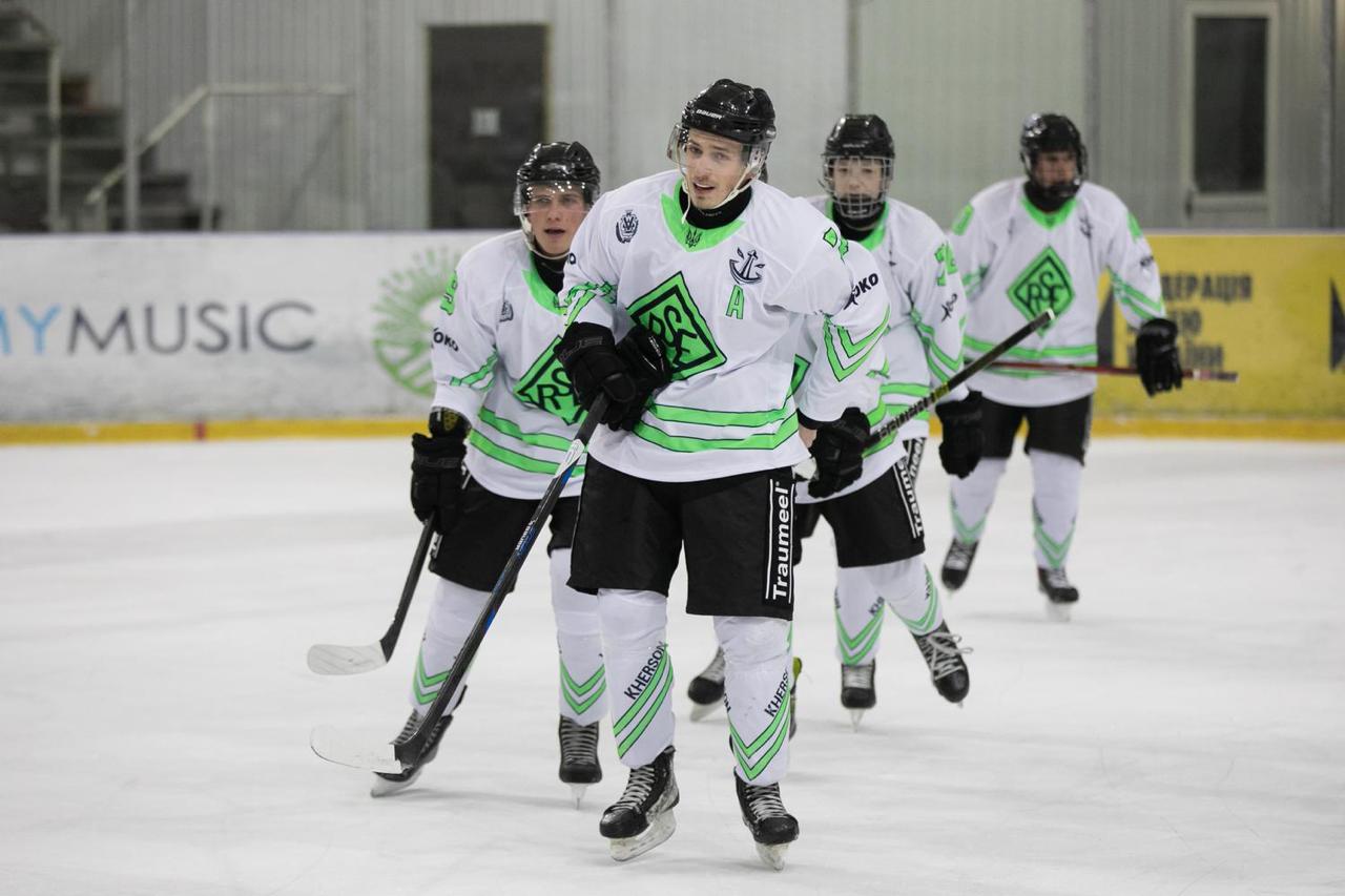 Чотири хокеїсти у білій формі з яскраво-зеленими смугами та логотипом на грудях ковзають по льодовій арені під час матчу. Гравці зосереджені, тримають ключки в руках, а на задньому плані видно захисний борт стадіону.