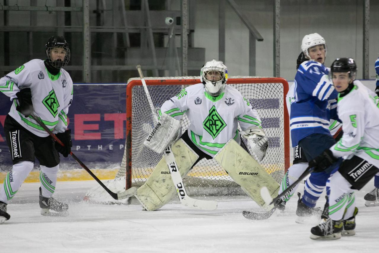 Напружений ігровий момент біля воріт під час хокейного матчу. Воротар у біло-зеленій формі стоїть у захисній стійці, готуючись відбити шайбу. Поруч перебувають гравці обох команд, ведеться активна боротьба на льодовому майданчику.