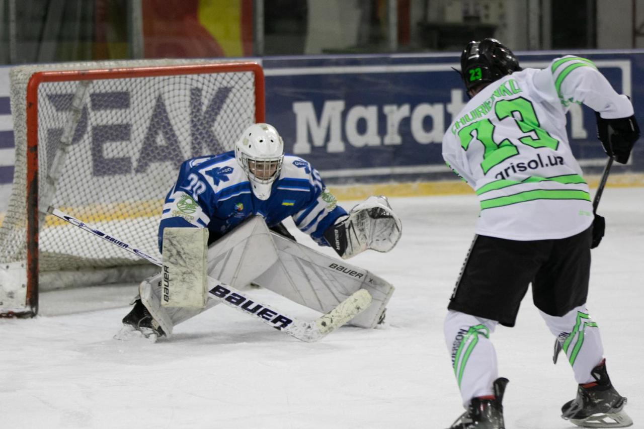 Голкіпер у синій формі надійно захищає ворота, перебуваючи у низькій стійці на льоду. Перед ним гравець суперника у біло-зеленому джерсі під номером 23 готується до атаки, тримаючи ключку. На задньому плані видно борт льодової арени з рекламними банерами.