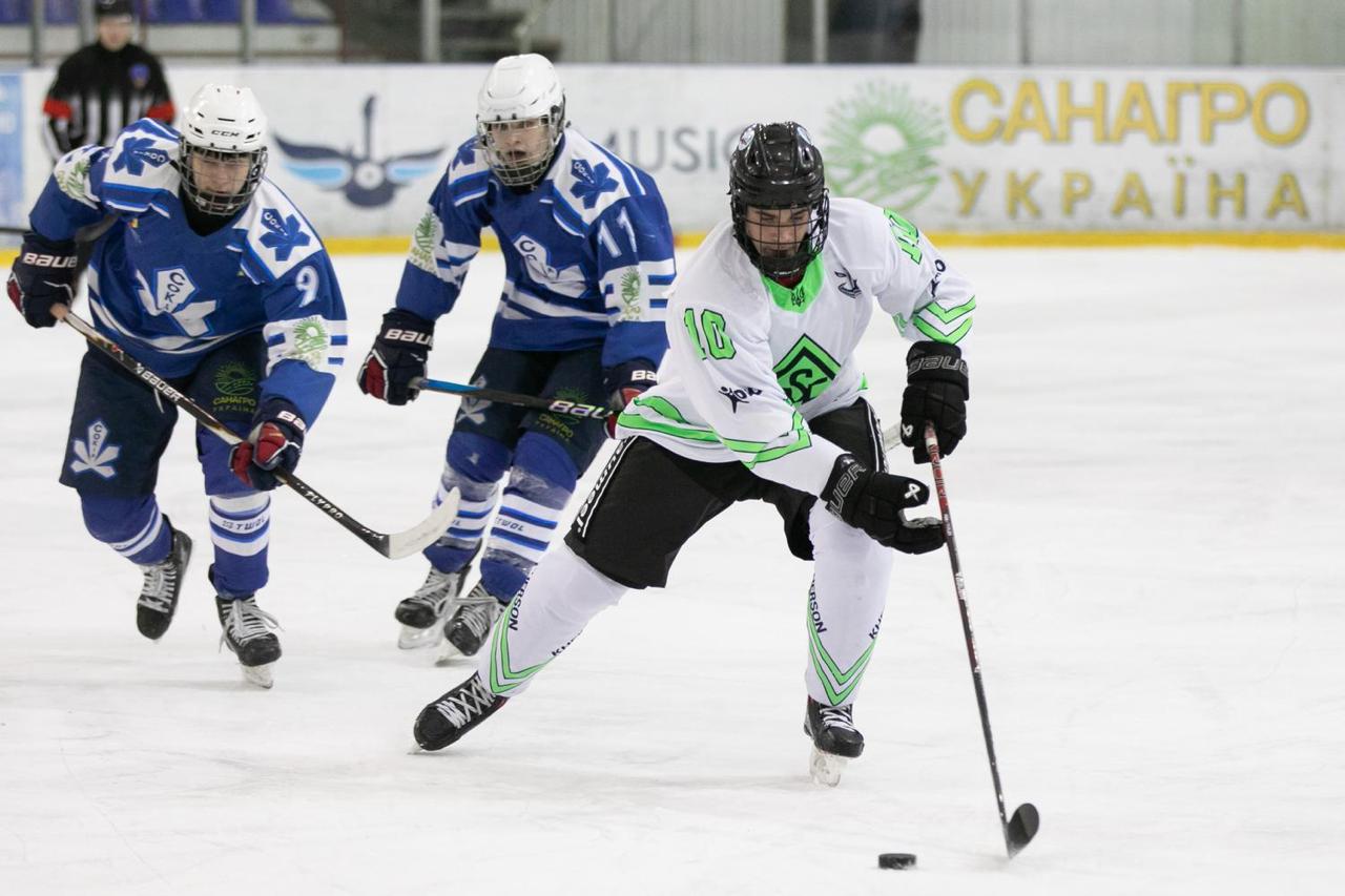 Нападник у білій формі з яскраво-зеленими елементами веде шайбу по льоду, готуючись до атаки. За ним слідують двоє захисників у синій формі, намагаючись перехопити ініціативу. На фоні льодової арени помітні рекламні банери з написами українською мовою.