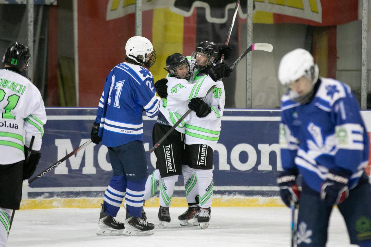 Двоє хокеїстів у білій формі з яскраво-зеленими елементами радісно обіймаються на льоду, святкуючи забиту шайбу. Поруч проїжджає гравець у синій формі, а на фоні видно захисний борт стадіону.