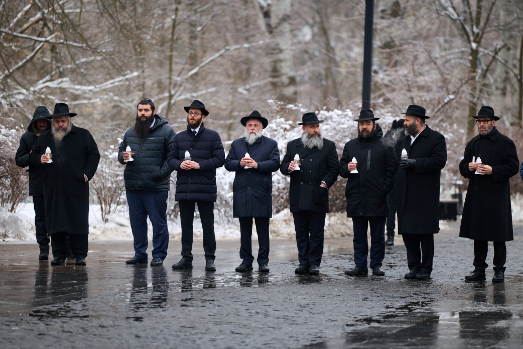 Група чоловіків у темному одязі та капелюхах стоїть на засніженій алеї. Кожен із них тримає в руках запалену білу лампадку, вшановуючи пам'ять під час меморіального заходу.