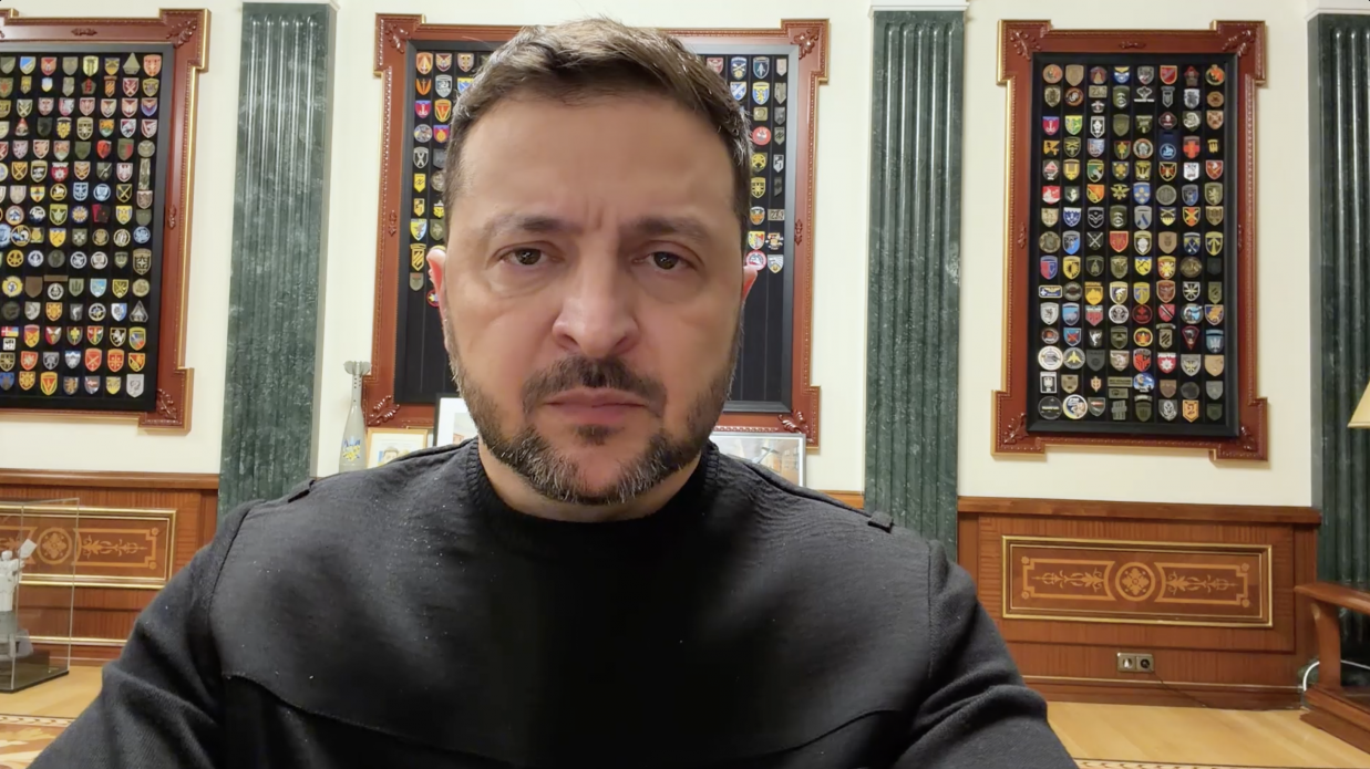 Чоловік у чорному светрі дивиться прямо в камеру на фоні офісного інтер’єру. Позаду нього на стінах розміщено три великі декоративні рамки з численними військовими шевронами та емблемами. Приміщення оздоблене дерев'яними панелями та зеленими колонами.