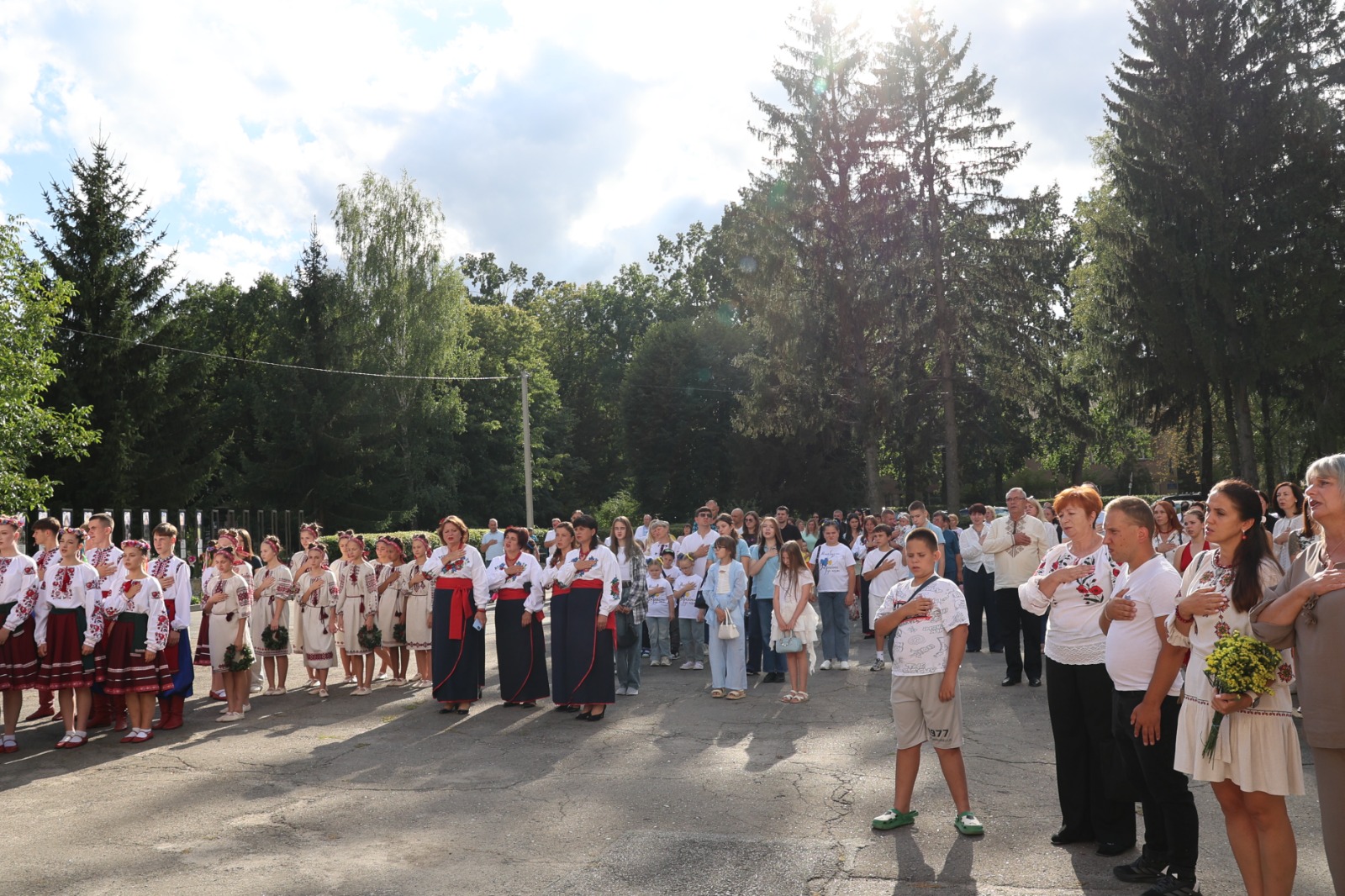 Мешканці громади та дитячі творчі колективи у вишиванках зібралися просто неба біля Алеї Слави для вшанування пам'яті захисників України. Більшість присутніх тримають праву руку на серці під час виконання Державного Гімну або хвилини мовчання. На задньому плані височіють зелені дерева під сонячним небом.