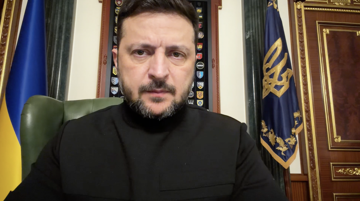 Чоловік у чорному одязі виступає зі зверненням на фоні державних символів України. Позаду нього з обох боків розміщені прапори, а на стіні по центру — панель із різними емблемами та шевронами.