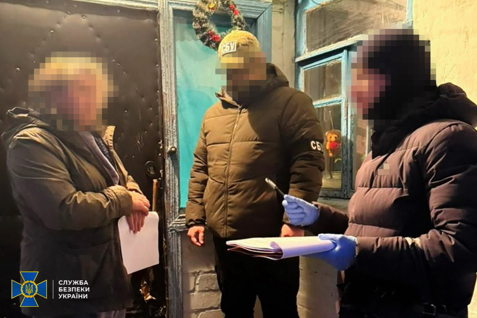 Троє осіб стоять біля входу в будинок. Співробітник СБУ в куртці кольору хакі та кепці з відповідним написом спостерігає за процесом. Інший правоохоронець у синіх рукавичках тримає документи та ручку, звертаючись до особи у цивільному одязі. Обличчя на фото розмиті для забезпечення анонімності. У нижньому куті розміщено логотип Служби безпеки України.