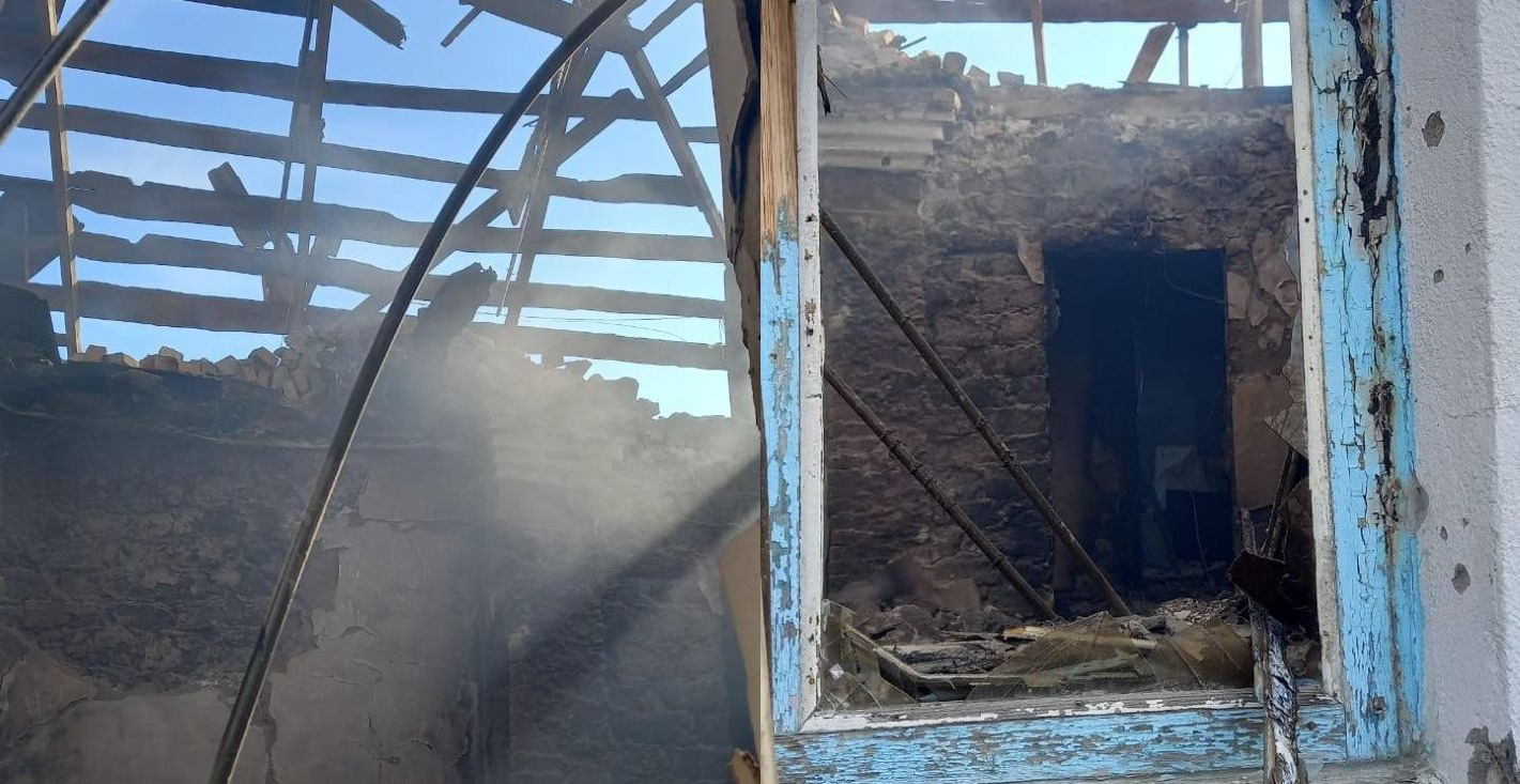 Зруйнована внаслідок обстрілу або пожежі будівля. На фото видно пошкоджений дах із оголеними дерев'яними балками, вибиті вікна та посічені уламками стіни. Крізь розбите вікно видно внутрішню частину цегляної стіни та задимлення.