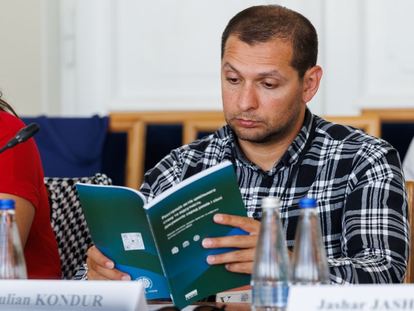 Радник Уповноваженого Офісу Омбудсмана України Юліан Кондур сидить за конференц-столом під час заходів БДІПЛ ОБСЄ у Варшаві та зосереджено вивчає «Збірник викликів, кращих практик та набутого досвіду в регіоні ОБСЄ: реєстрація актів цивільного стану та відсутність документів серед ромів і сінті». Чоловік у сорочці в чорно-білу клітинку зафіксований по центру кадру великим планом, схиливши голову над бірюзово-зеленою обкладинкою друкованого видання. Перед ним на столі розставлені скляні пляшки з мінеральною водою, а ліворуч на передньому плані розташована його іменна табличка «Julian KONDUR». Засідання присвячене питанням подолання бездокументованості та реалізації права на юридичну ідентичність національних меншин.