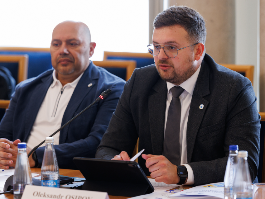 Представник Уповноваженого Офісу Омбудсмана України Олександр Осіпов в окулярах та діловому костюмі виступає на заходах БДІПЛ ОБСЄ у Варшаві, присвячених захисту прав ромів та сінті. Посадовець сидить праворуч за конференц-столом перед планшетом у чорному чохлі та паперами, тримаючи в руці ручку під час представлення напрацювань щодо додержання прав національних меншин. Ліворуч від нього за столом розмістився начальник відділу з питань захисту прав осіб з числа ромської національної меншини Володимир Кондур у темно-синьому піджаці поверх білої сорочки-поло, який уважно слухає виступ колеги. На столі перед учасниками також розставлені пляшки з мінеральною водою та встановлено настільні мікрофони.