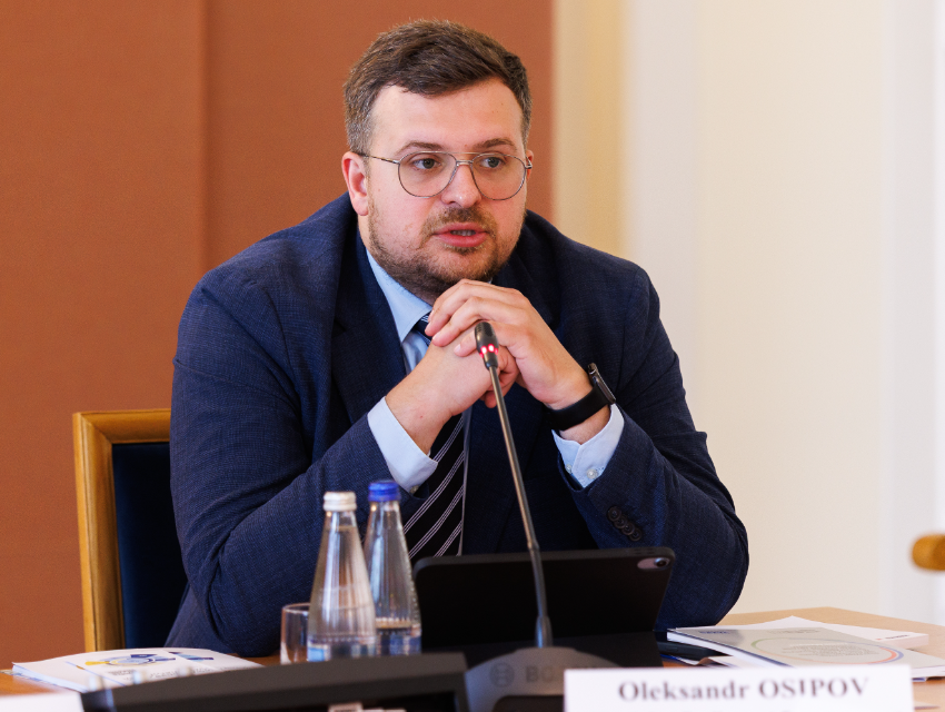 Представник Уповноваженого Офісу Омбудсмана України Олександр Осіпов в окулярах та діловому костюмі виступає на заходах БДІПЛ ОБСЄ у Варшаві, присвячених захисту прав ромів та сінті. Посадовець зафіксований по центру кадру великим планом, сидить за дерев'яним столом, зчепивши пальці рук перед собою біля настільного мікрофона під час представлення напрацювань Офісу. Він одягнений у темно-синій піджак, світло-блакитну сорочку та краватку у смужку. Перед ним лежать документи й відкритий планшет у чорному чохлі, ліворуч стоять пляшки з водою, а на передньому плані розташована його іменна табличка «Oleksandr OSIPOV».