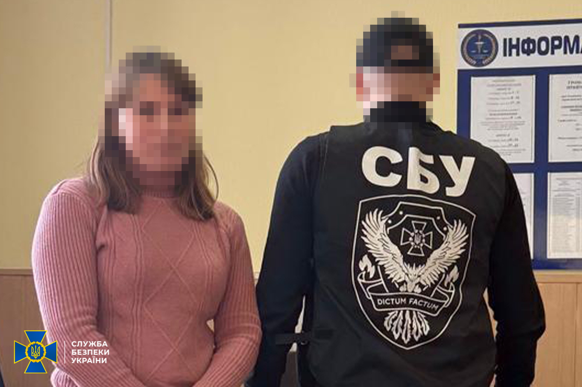 Жінка у рожевому светрі стоїть поруч із правоохоронцем у жилеті з написом «СБУ» на тлі інформаційного стенда. Обличчя на фотографії розмиті для збереження анонімності. У нижньому лівому куті розміщено логотип Служби безпеки України.