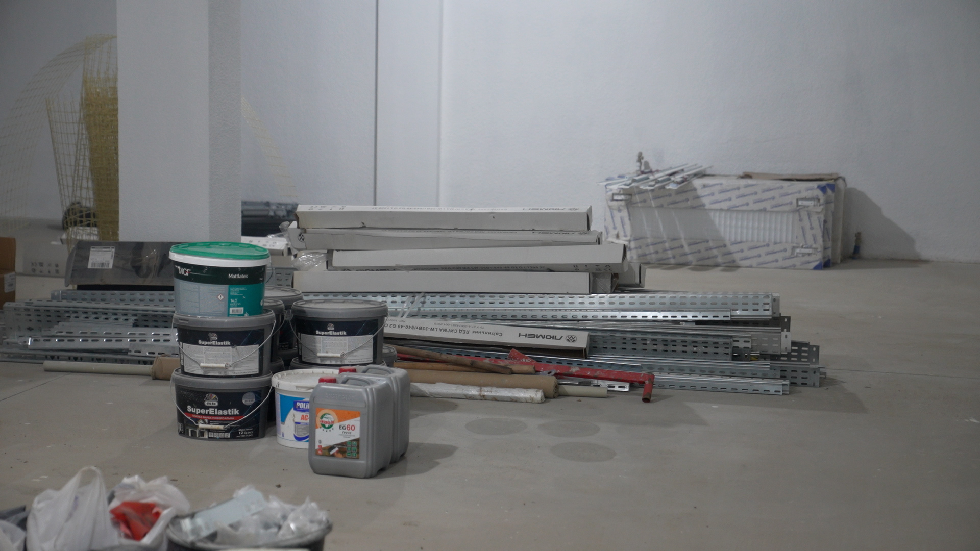 Будівельні матеріали, складені на підлозі в приміщенні з білими стінами. На передньому плані видно відра з фарбою та ґрунтовкою, каністри, металеві профілі для гіпсокартону та довгі картонні коробки.