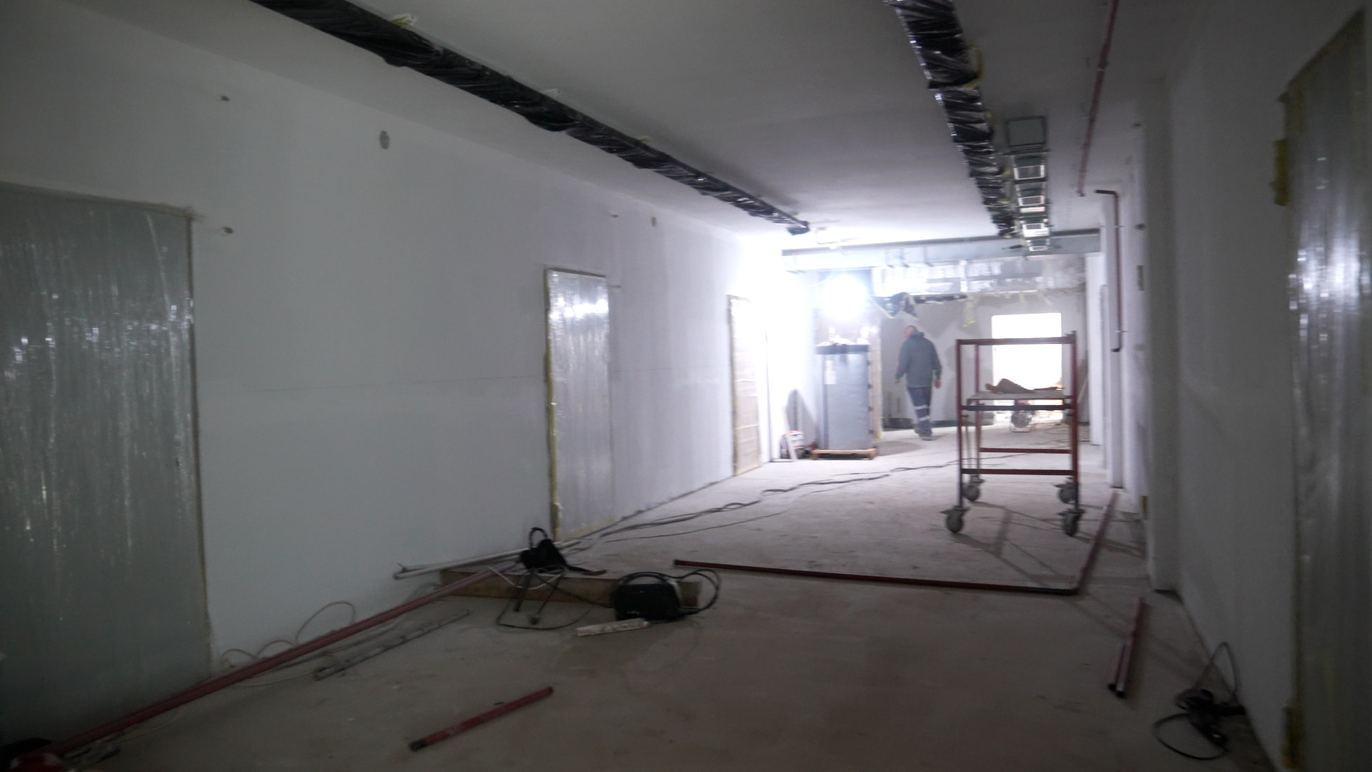 Інтер'єр довгого коридору на стадії капітального ремонту. Стіни поштукатурені та пофарбовані у білий колір, двері захищені плівкою, на стелі змонтовані комунікації, а вдалині видно будівельне риштування та силует робітника.