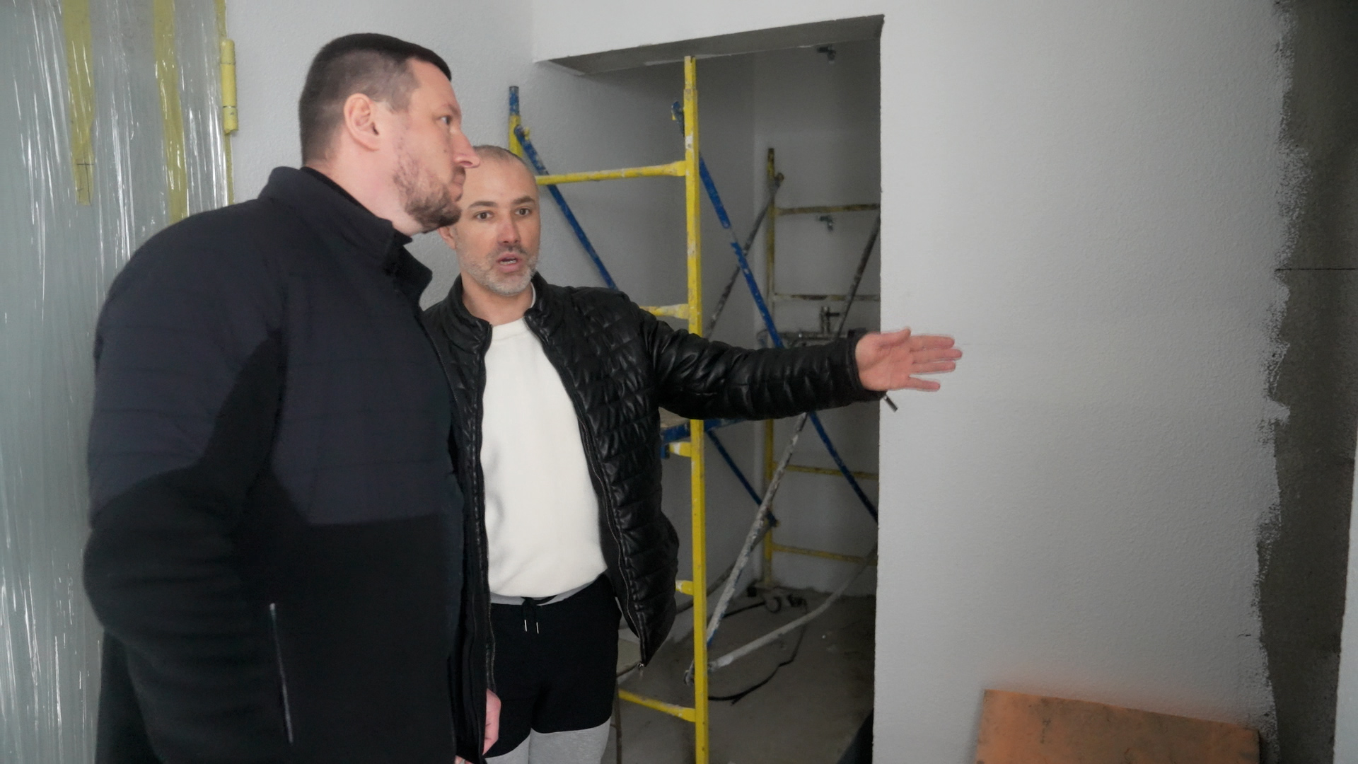 Двоє чоловіків у верхньому одязі оглядають приміщення під час ремонту. Один із них вказує рукою на стіну, обговорюючи робочий процес. На задньому плані видно жовте будівельне риштування у дверному отворі.