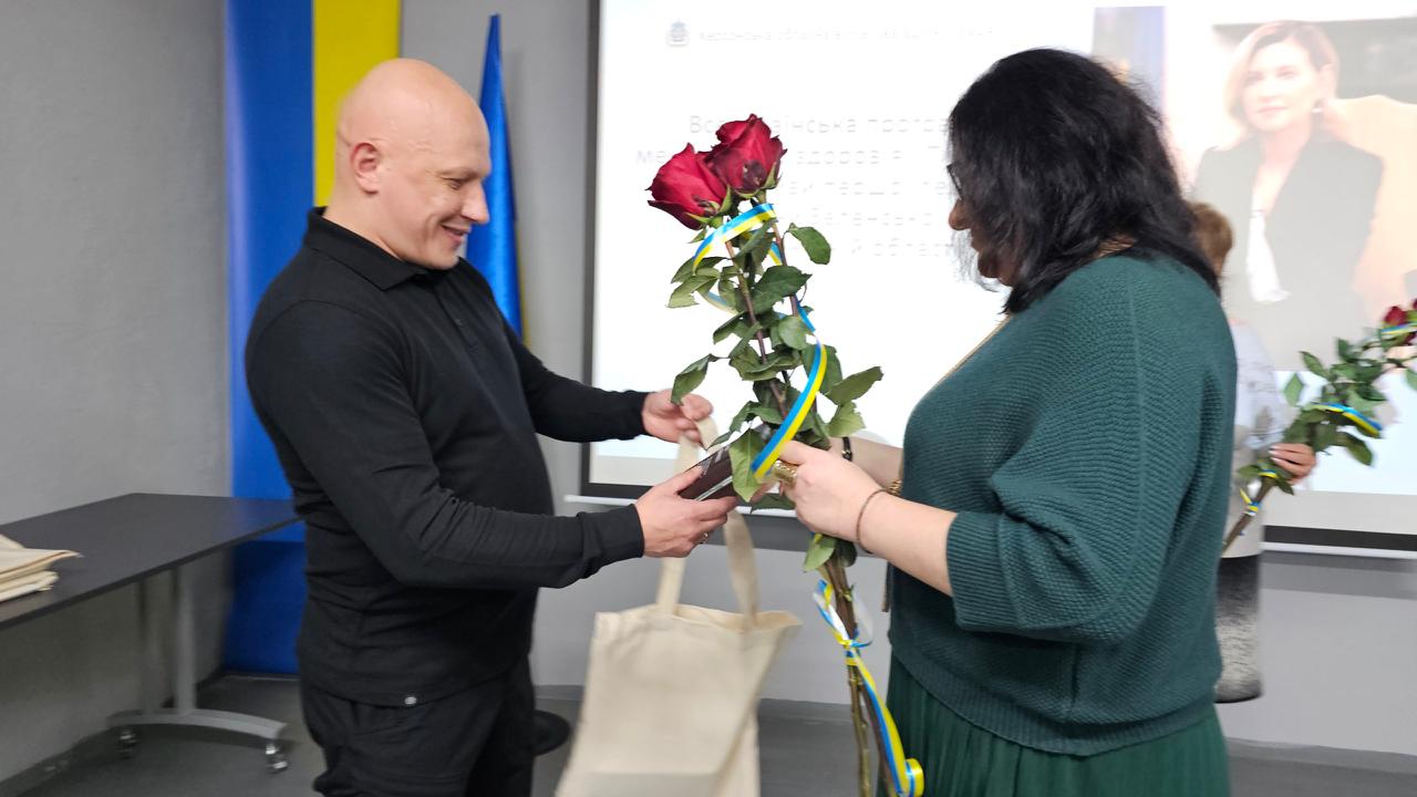 Чоловік у чорній сорочці посміхається, вручаючи жінці, одягненій у темно-зелений светр, букет червоних троянд, прикрашених синьо-жовтими стрічками. На задньому плані видно екран з текстом про Всеукраїнську програму ментального здоров'я.