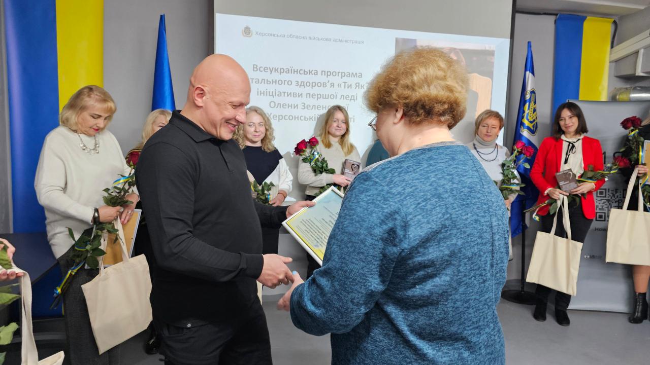 Чоловік у чорній сорочці посміхається, вручаючи нагородний документ жінці в синьому светрі під час офіційного заходу. На задньому плані стоять інші жінки з букетами троянд, а на екрані відображається інформація про Всеукраїнську програму ментального здоров'я Ти як? у Херсонській області.