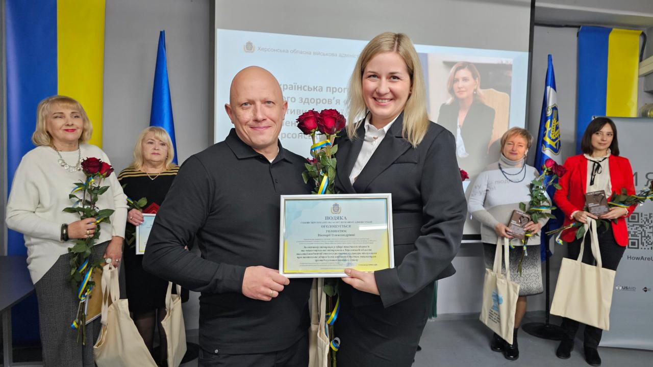 Чоловік у чорній сорочці та жінка в чорному діловому костюмі стоять разом, тримаючи в руках подяку, обрамлену синьо-жовтими кольорами, та букет червоних троянд. Обидва посміхаються. На задньому плані видно екран із логотипом Херсонської ОВА та фотографією Олени Зеленської, а також інших учасників заходу.
