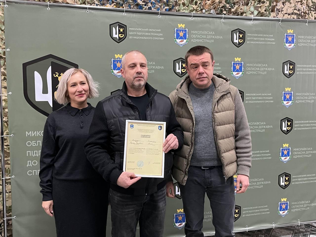 Троє людей (двоє чоловіків та жінка) стоять перед стіною з емблемами та написами: Миколаївська обласна державна адміністрація. Чоловік посередині, одягнений у чорний жилет, тримає в руках рамку із документом про проходження базової військової підготовки.