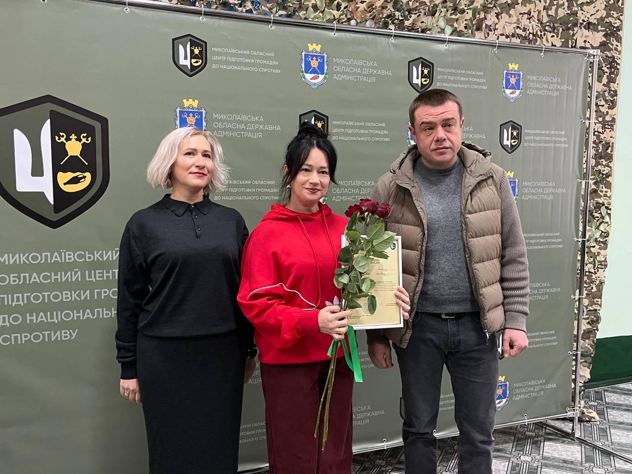 Груповий знімок трьох осіб (двоє жінок і чоловік) на тлі банера Миколаївської обласної державної адміністрації. Жінка посередині, одягнена в червоний одяг, тримає букет червоних троянд та документ про проходження базової військової підготовки.