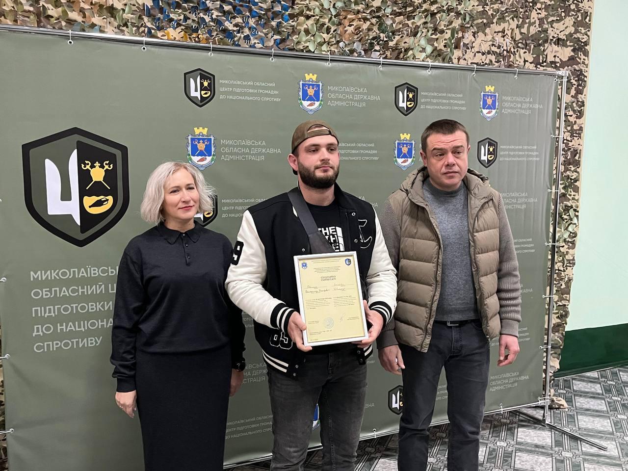 Груповий знімок жінки та двох чоловіків, що стоять перед фоном з емблемами та написами: Миколаївська обласна державна адміністрація. Молодий чоловік у кепці та світло-чорній куртці, який стоїть посередині, тримає в руках рамку із документом про проходження базової військової підготовки.