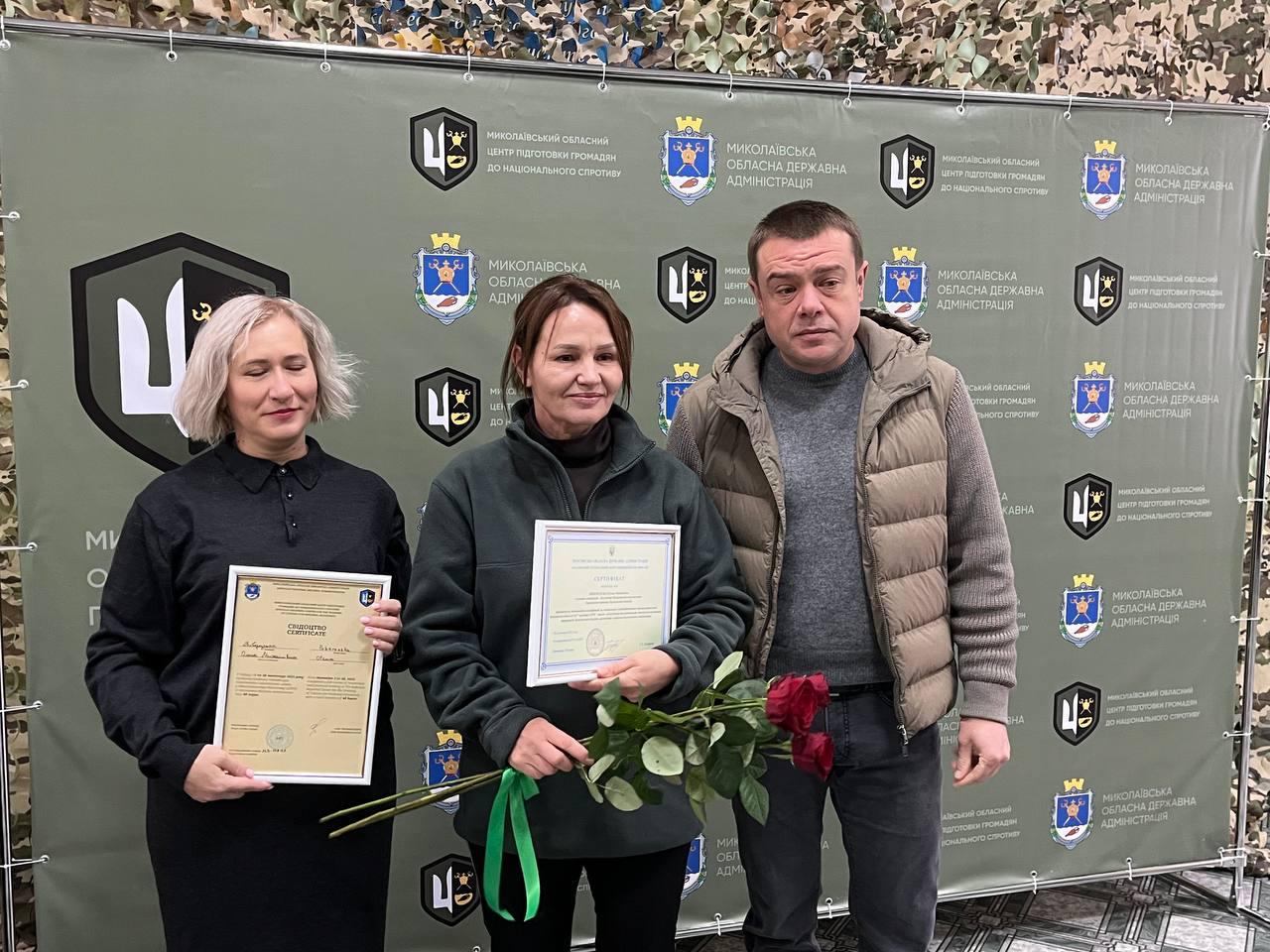 Групове фото трьох осіб (двоє жінок та чоловік) на тлі банера Миколаївської обласної державної адміністрації. Жінки тримають документи про проходження базової військової підготовки, а жінка посередині також тримає букет червоних троянд.