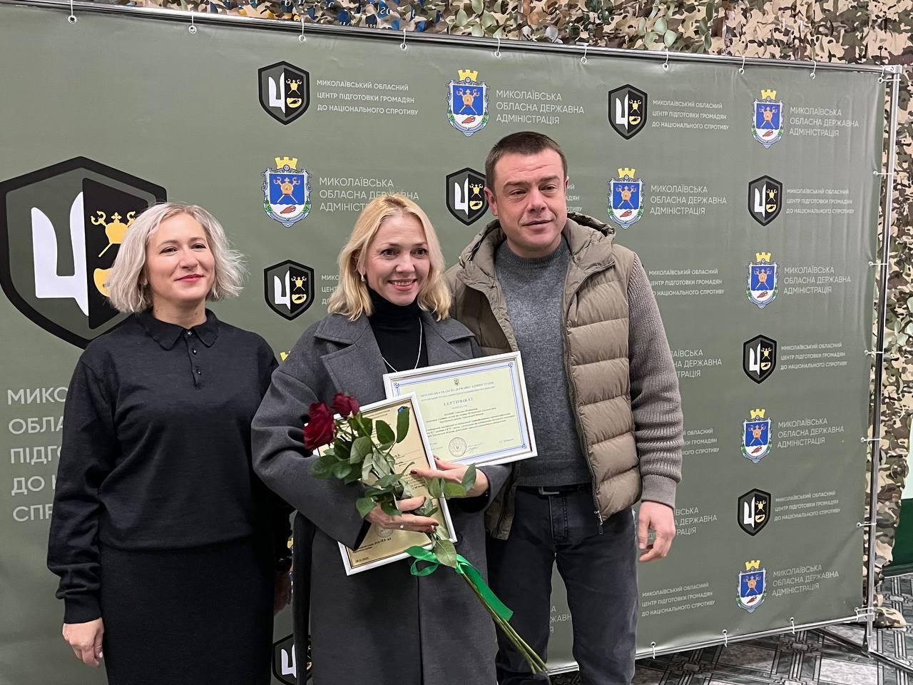 Груповий знімок трьох осіб (двох жінок і чоловіка) на офіційному заході. Жінка посередині у сірому пальті посміхається, тримаючи букет червоних троянд та два документи про проходження базової військової підготовки на тлі банера Миколаївської обласної державної адміністрації.