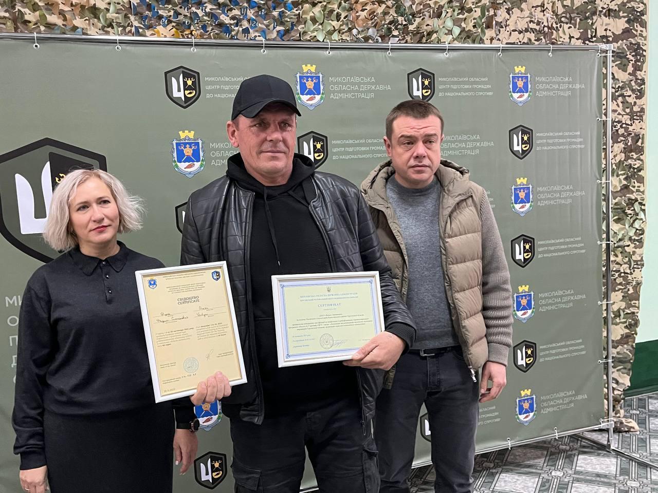 Груповий знімок трьох осіб (жінки та двох чоловіків) на тлі банера Миколаївської обласної державної адміністрації. Чоловік посередині в чорній шкіряній куртці та кепці тримає два документи про проходження базової військової підготовки.