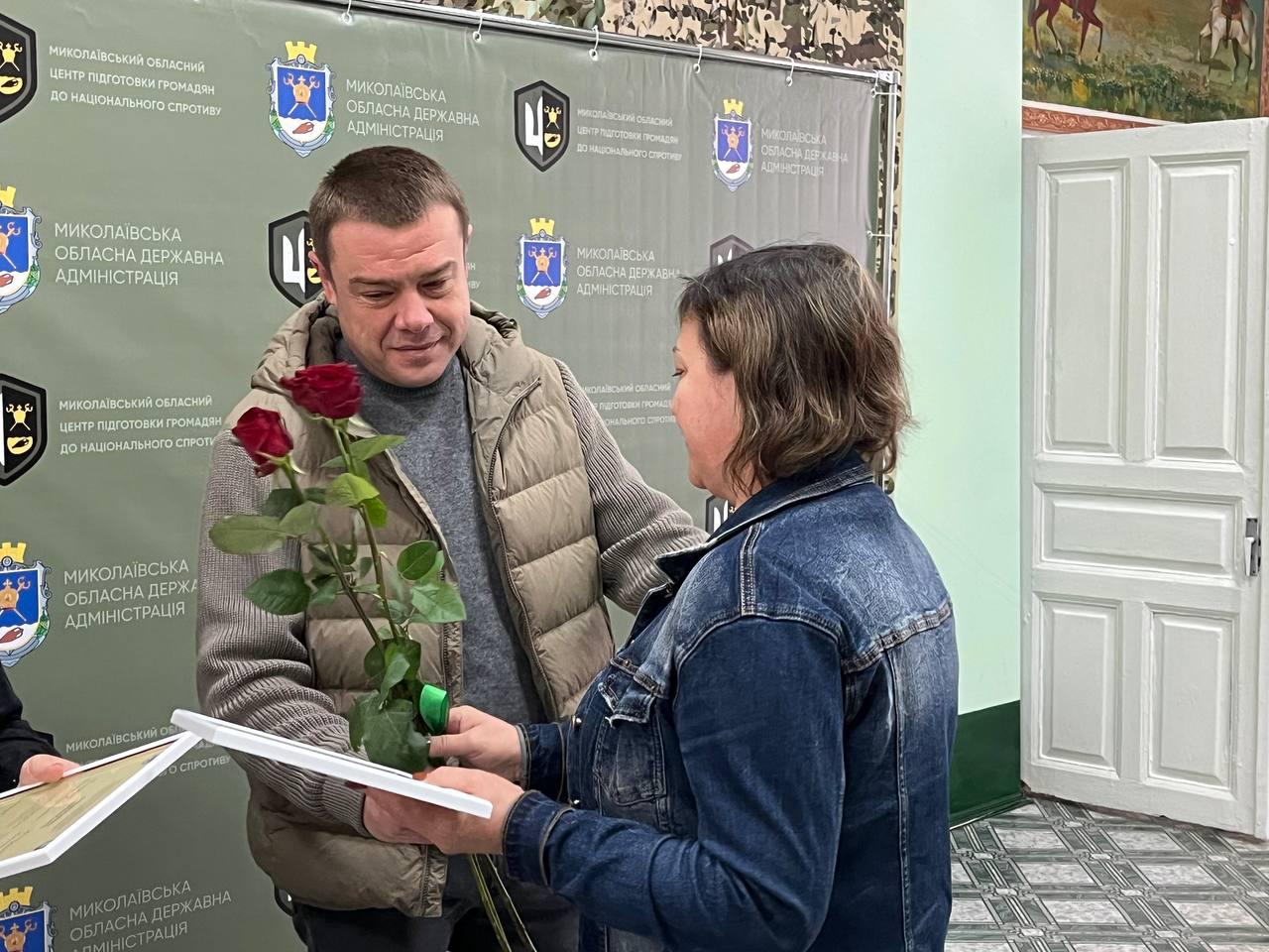 Двоє людей, чоловік у жилетці та жінка в джинсовій куртці, стоять обличчям одне до одного на тлі банера Миколаївської обласної державної адміністрації. Жінка передає чоловікові букет червоних троянд і документ про проходження базової військової підготовки.
