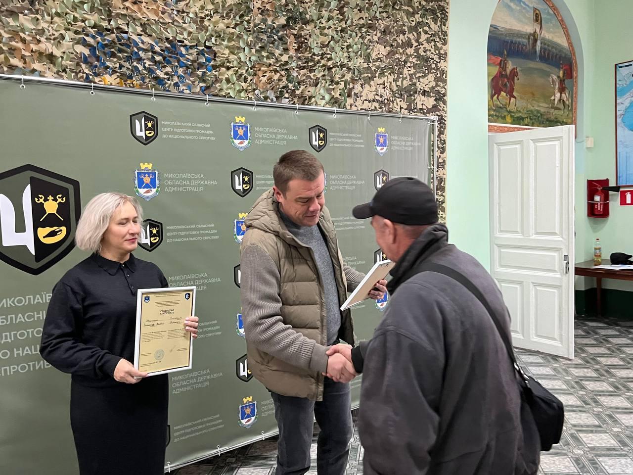 Чоловік у жилетці тисне руку літньому чоловікові в кепці та темному одязі, обмінюючись документами. Ліворуч стоїть жінка, яка тримає в руках документ про проходження базової військової підготовки. Подія відбувається на тлі банера Миколаївської обласної державної адміністрації, у приміщенні із картиною на стіні.