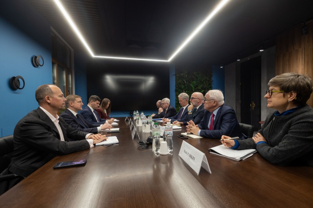 Група офіційних осіб у ділових костюмах сидить за довгим столом у сучасній конференц-залі з синіми стінами та лінійним освітленням на стелі. На столі розміщені таблички з іменами учасників, блокноти для записів та пляшки з водою. Триває робоча зустріч або переговори.