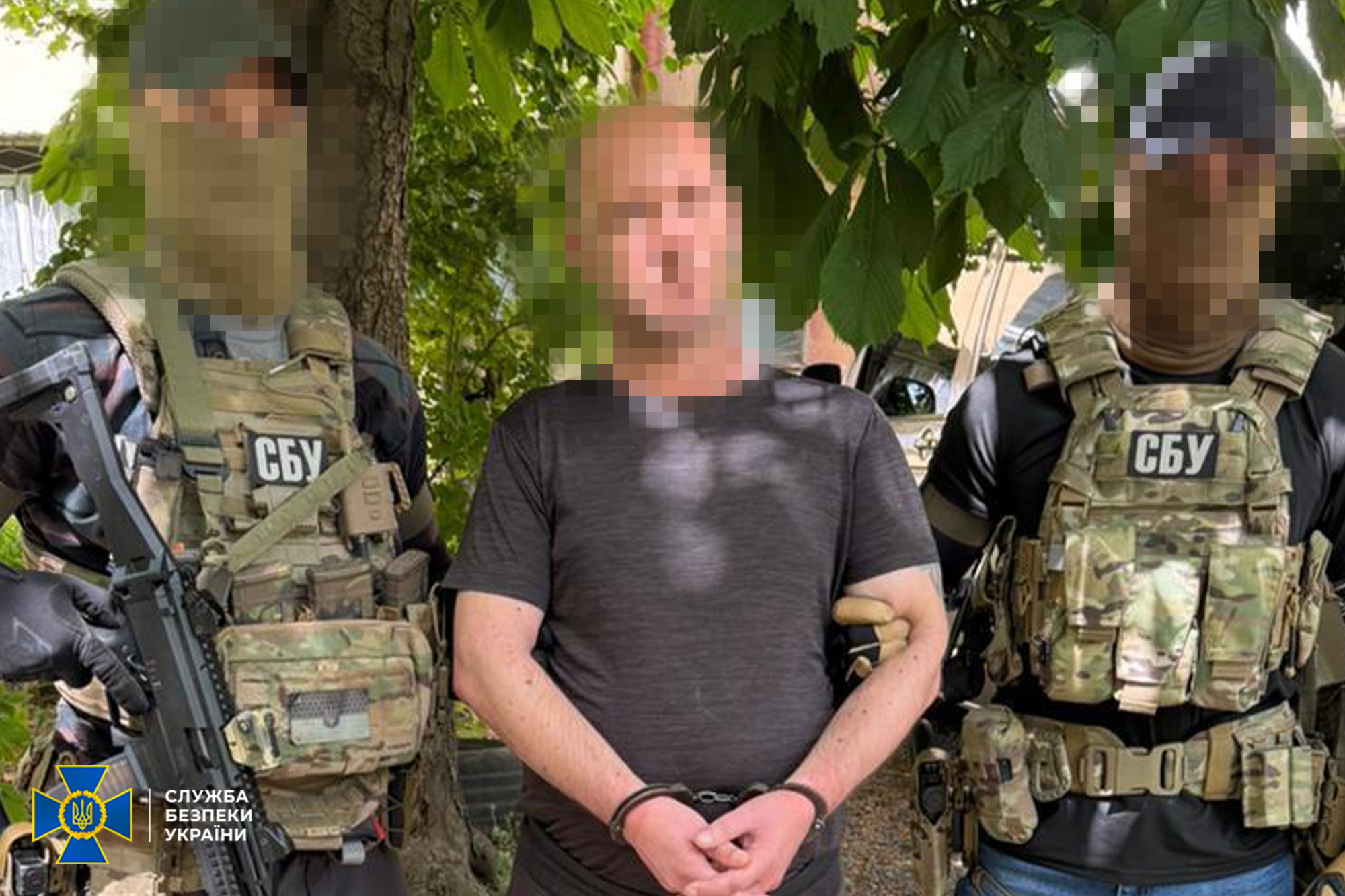 Затримання підозрюваного співробітниками Служби безпеки України. По центру стоїть затриманий чоловік у темно-сірій футболці, його руки спереду закованi в наручники. По обидва боки від нього стоять двоє правоохоронців у повній бойовій екіпіровці — камуфляжних тактичних жилетах із розвантаженнями, балаклавах та захисних окулярах. На жилетах військових чітко видно нашивки з написом «СБУ». Силовик ліворуч тримає в руках автоматичну зброю. Затримання відбувається на вулиці на тлі зелених дерев, обличчя всіх трьох чоловіків розмиті за допомогою піксельного фільтра. У лівому нижньому кутку розміщено офіційний логотип СБУ із написом «СЛУЖБА БЕЗПЕКИ УКРАЇНИ».