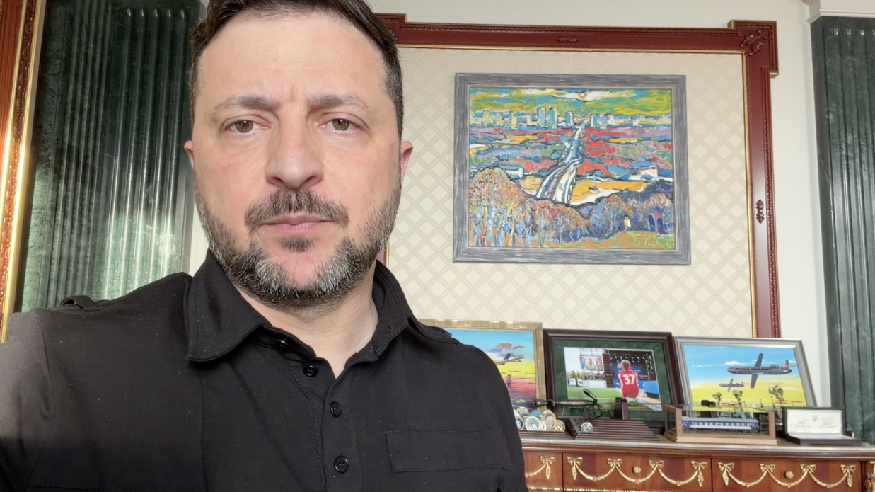Президент України Володимир Зеленський під час офіційного відеозвернення. Він зображений великим планом на передньому плані ліворуч, одягнений у чорну сорочку поло. Його погляд зосереджений та спрямований прямо в камеру.  На задньому плані — стіна кабінету зі світлими шпалерами у ромбоподібний візерунок та великою дерев'яною багетною рамою. Всередині рами висить яскрава картина у сірому канті, що зображує міський пейзаж та річку або дорогу в імпресіоністичному стилі. Під картиною на дерев'яному комоді з позолоченим оздобленням розставлені особисті речі: три невеликі малюнки у рамках (на одному зображено літак у небі, на іншому — футболіст у червоній формі з номером 37), колекційні монети, макет потяга у скляному футлярі та інші пам'ятні сувеніри. По боках кадру видно темно-зелені інтер'єрні колони.
