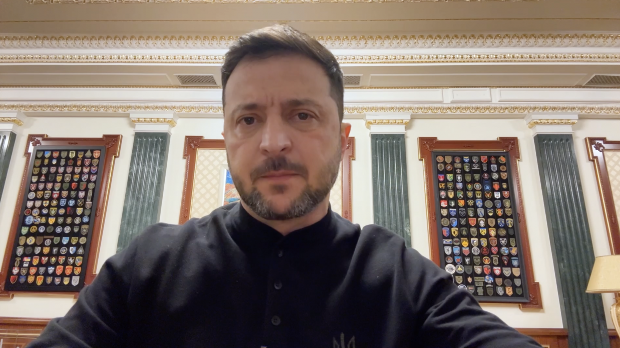 Президент України Володимир Зеленський у чорній сорочці, з бородою, дивиться прямо в камеру. Він стоїть у приміщенні з вишуканим оздобленням, а за ним на стіні висять дві великі вітрини з колекціями нашивок і знаків розрізнення різних українських військових підрозділів.