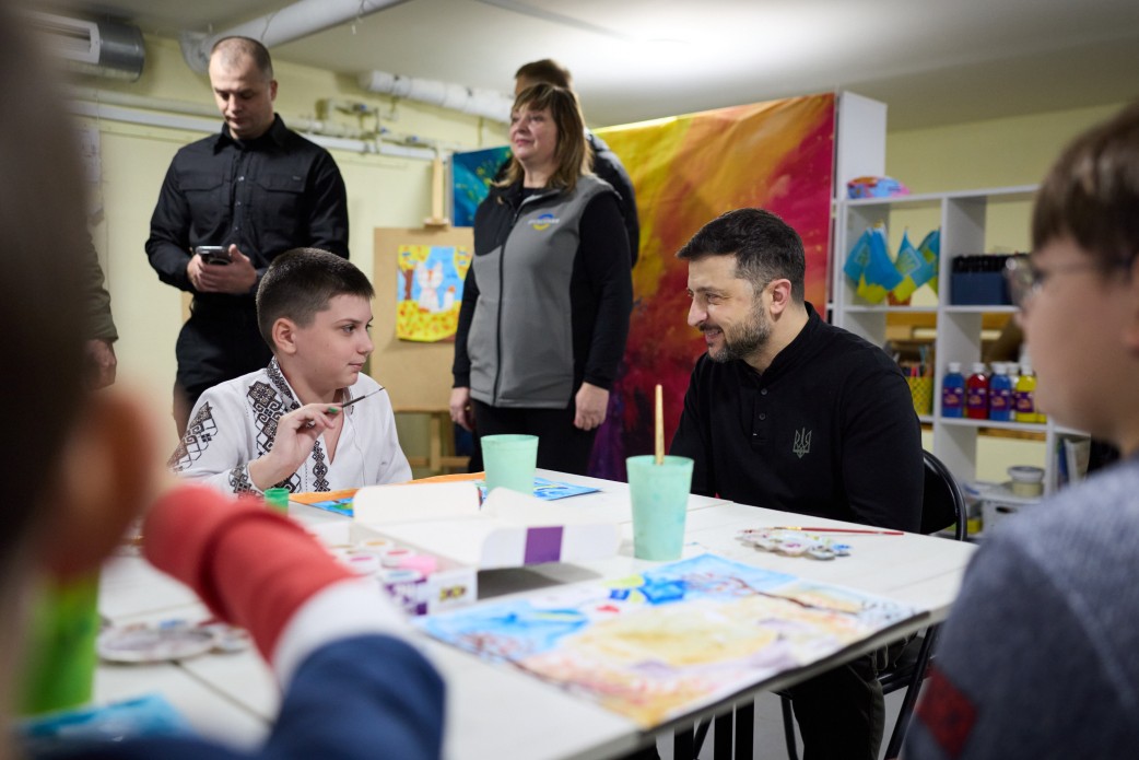 Президент України Володимир Зеленський (в чорній кофті) сидить за столом і розмовляє з хлопчиком у вишиванці, який тримає пензель. Вони знаходяться в приміщенні, де проводяться заняття з малювання. На задньому плані стоять дорослі.