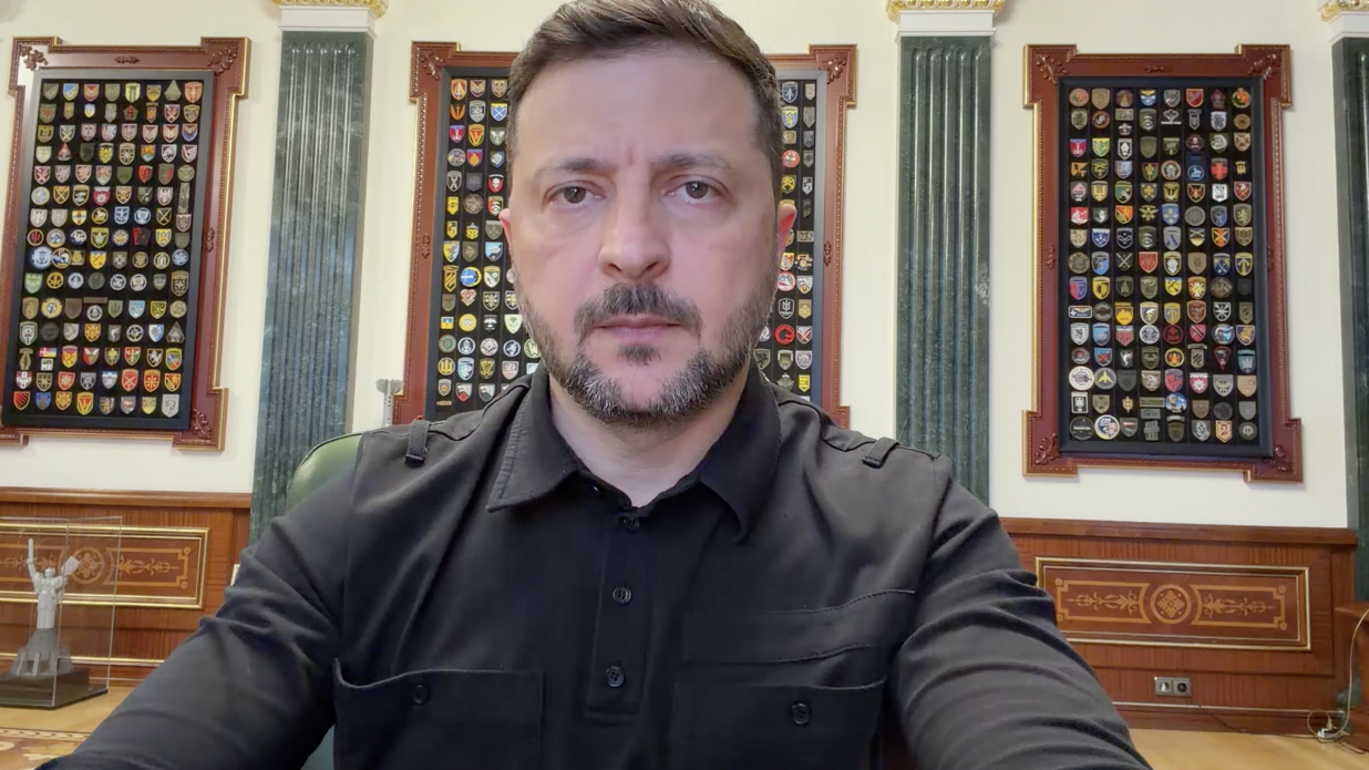 Президент України Володимир Зеленський під час офіційного відеозвернення. Він зображений великим планом, одягнений у чорну тактичну сорочку поло з довгим рукавом та накладними кишенями. На задньому плані — інтер'єр робочого кабінету з різьбленими дерев'яними панелями, зеленими мармуровими колонами та трьома великими вертикальними стендами, заповненими шевронами різних підрозділів Сил оборони України. Погляд Президента серйозний та зосереджений, спрямований прямо в камеру.