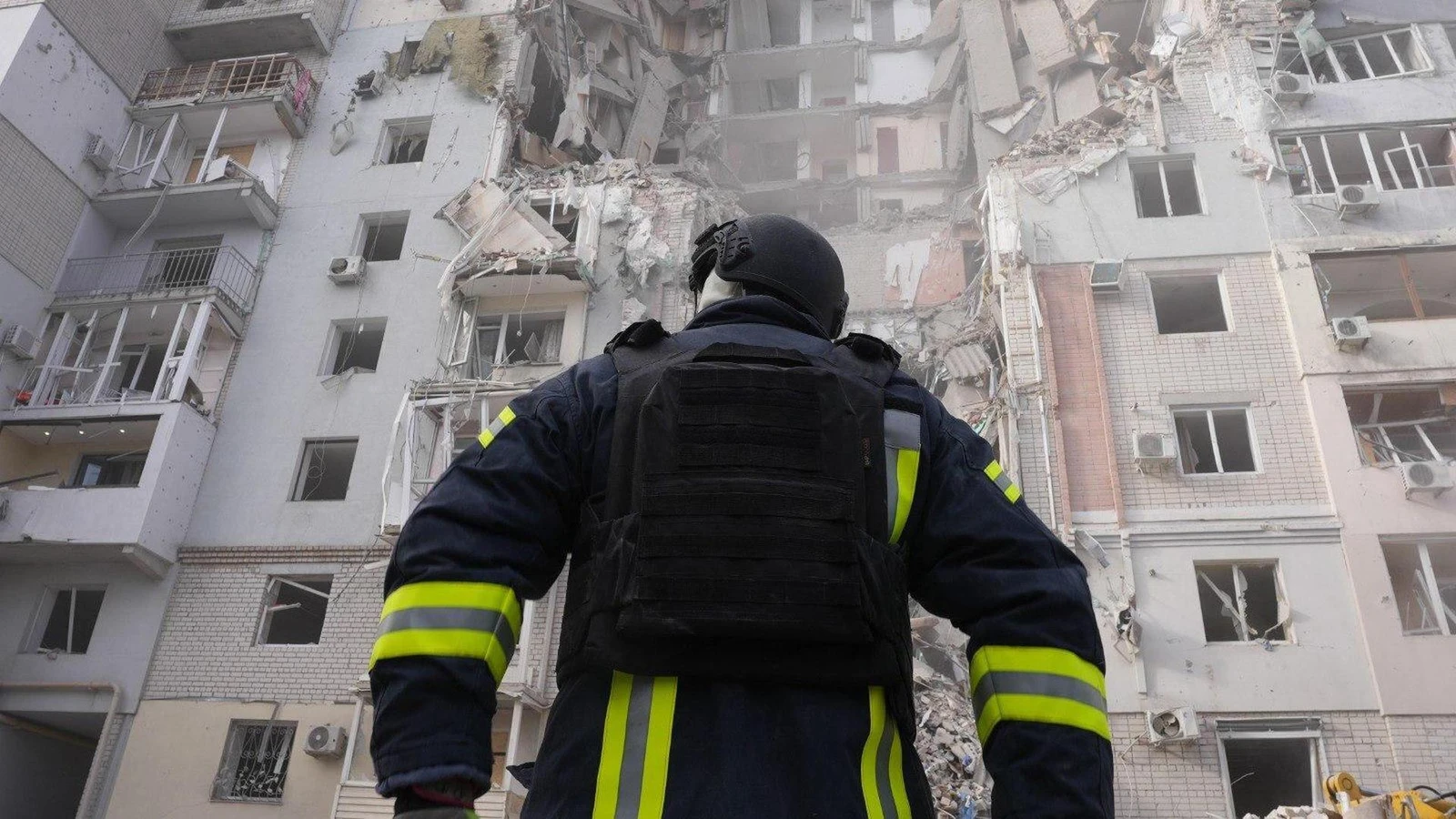 Рятувальник ДСНС стоїть спиною до камери на фоні багатоповерхового житлового будинку, значно пошкодженого внаслідок обстрілу. Видно зруйновані стіни, вибиті вікна та бетонні перекриття будівлі.