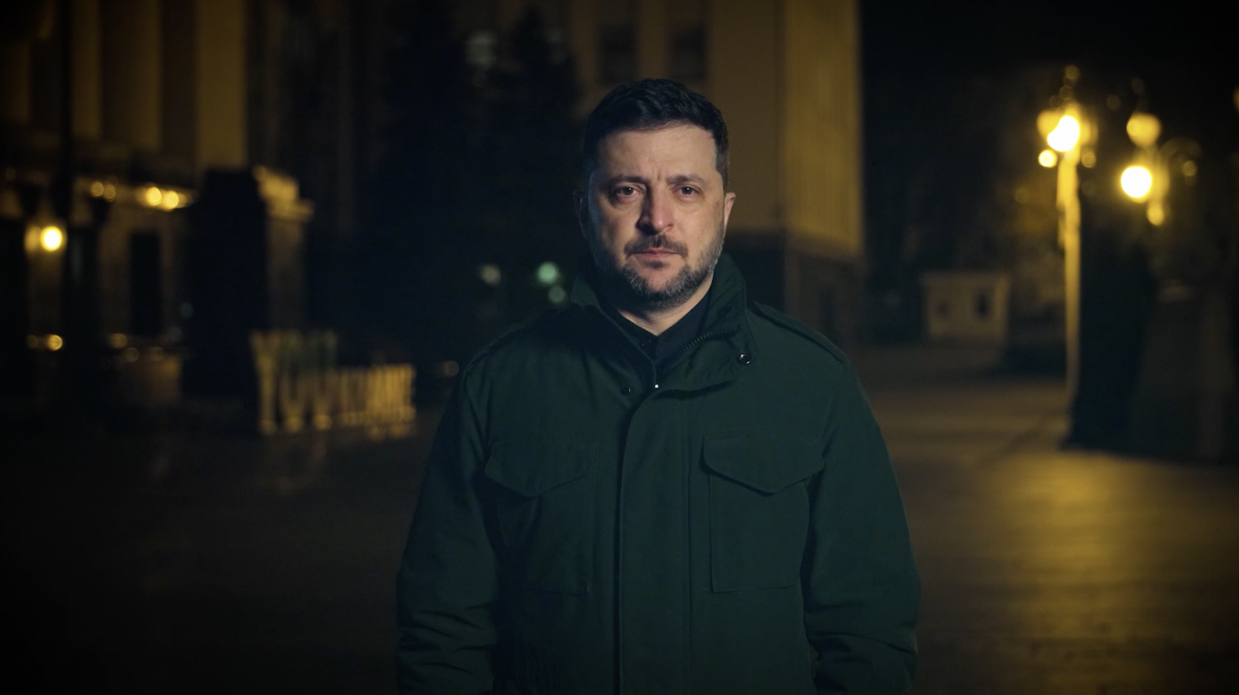 Президент України Володимир Зеленський стоїть на вулиці в темно-зеленій куртці вночі, дивлячись прямо в камеру. За ним видно освітлені будівлі та вуличні ліхтарі.