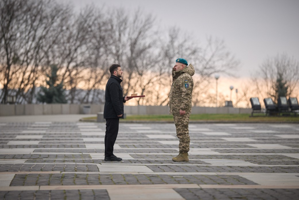 На фото зображений Президент України Володимир Зеленський у чорній куртці, який вручає нагороду військовослужбовцю в камуфляжній формі та темно-синьому береті. Президент тримає нагороду на червоному лотку. Церемонія відбувається на вулиці, на задньому плані стоять інші військові та цивільні особи.