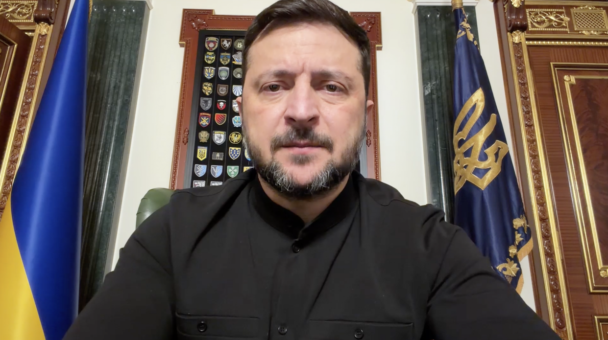 Чоловік середнього віку з темним волоссям і бородою, одягнений у чорну сорочку, дивиться прямо в камеру на фоні державних прапорів та офіційного інтер'єру.