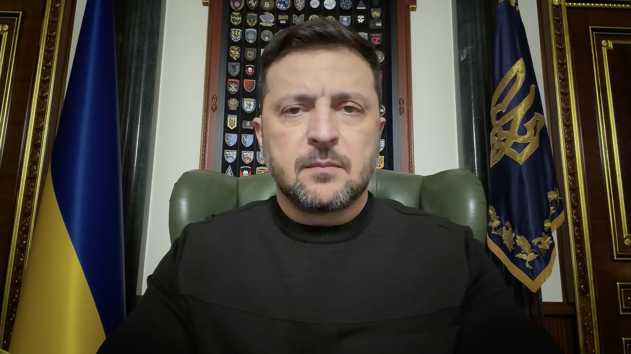 Чоловік у чорному светрі сидить у кабінеті між державним прапором та стягом із гербом. Позаду нього розміщена колекція військових шевронів у рамці.