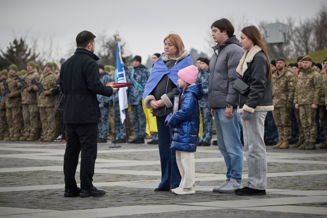 На фото зображений Президент України Володимир Зеленський у чорній куртці, який стоїть перед сім'єю (жінкою, хлопцем, дівчиною-підлітком та дівчинкою) щоб вручити нагороду родині загиблого військовослужбовця. Жінка накрита прапором України. На задньому плані стоять військові в строю.