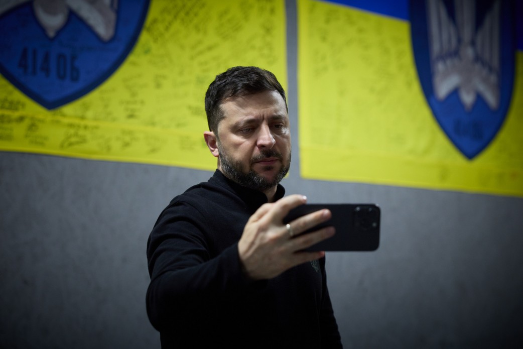 Чоловік із темною бородою, одягнений у чорну водолазку, а саме Президент України Володимир Зеленський, дивиться на екран мобільного телефону, який тримає у руці. На задньому плані видно два жовто-сині прапори з великими гербами та написами, ймовірно, військових підрозділів.
