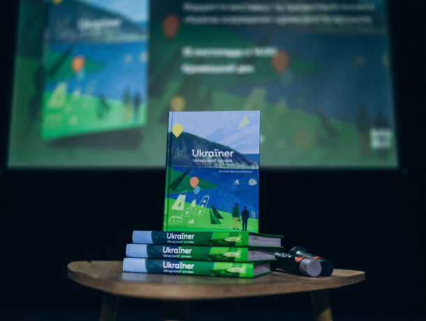 На дерев’яному столику лежить стос книг «Ukraïner», одна з яких стоїть вертикально. На задньому плані — розмитий екран із презентацією цієї ж книги.