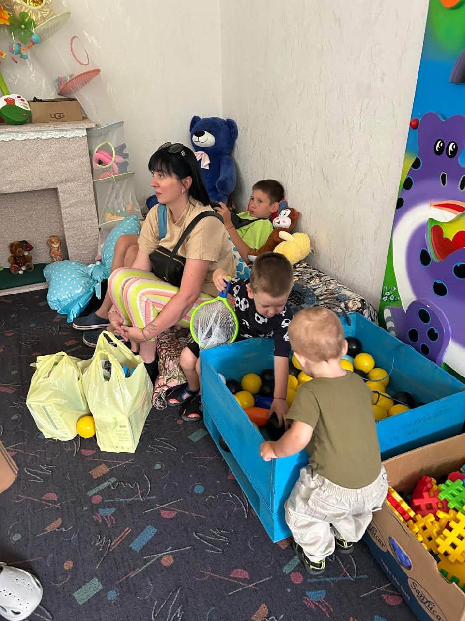 Жінка та кілька дітей у дитячій ігровій кімнаті. Жінка в бежевій футболці та смугастих легінсах сидить на дивані біля кутка кімнати поруч із м'якими іграшками та подушками, спостерігаючи за дітьми. Перед нею на підлозі стоять жовті пакети з покупками або допомогою. Двоє маленьких хлопчиків граються біля великої синьої дерев'яної коробки, наповненої жовтими та чорними пластиковими кульками, а поруч на килимі стоїть коробка з кольоровим конструктором.
